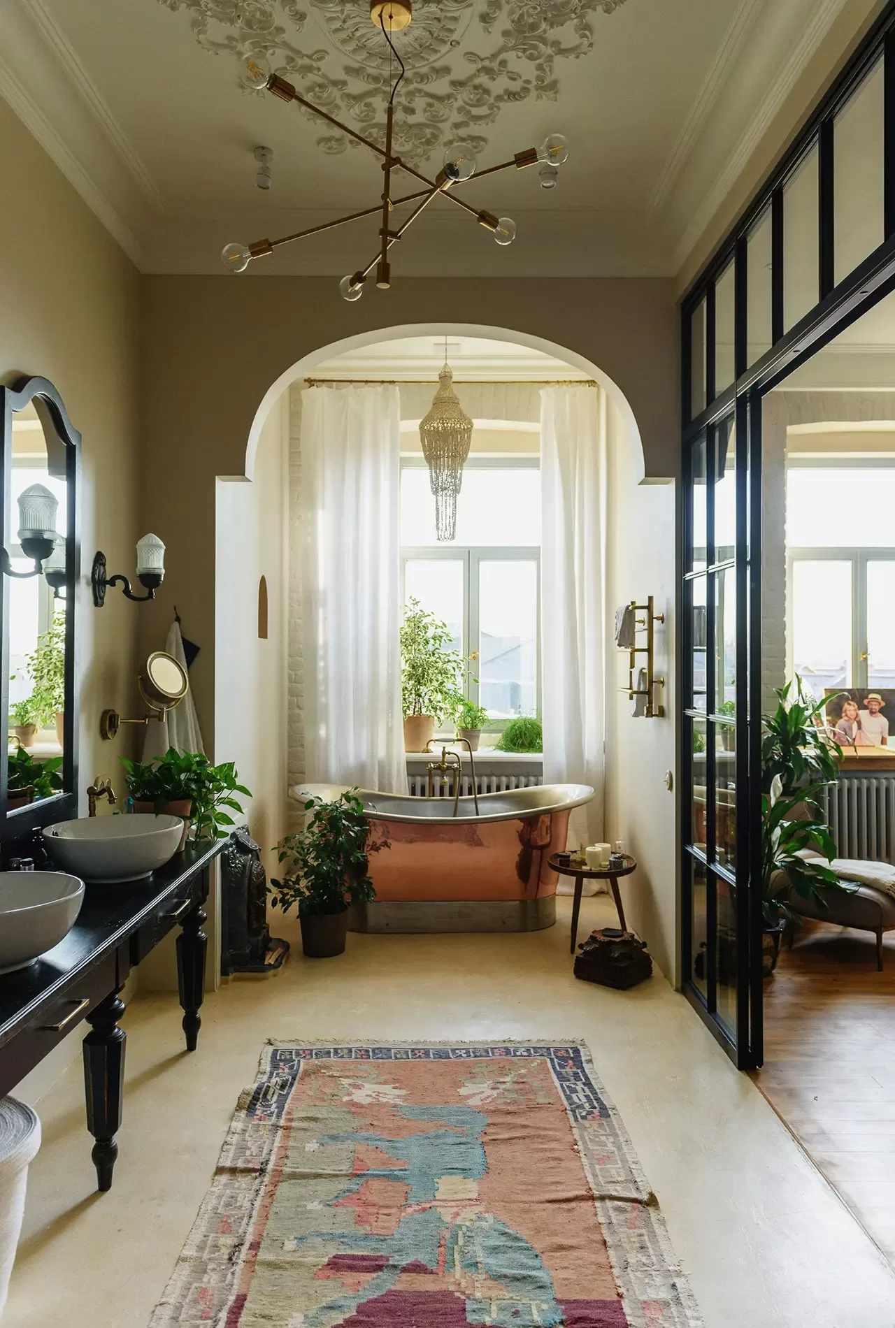 Elegant bathroom with a freestanding bathtub under an arch and chandelier, surrounded by plants. Two sinks stand on a black counter, and a colorful rug lies on the floor. Large windows let in natural light.