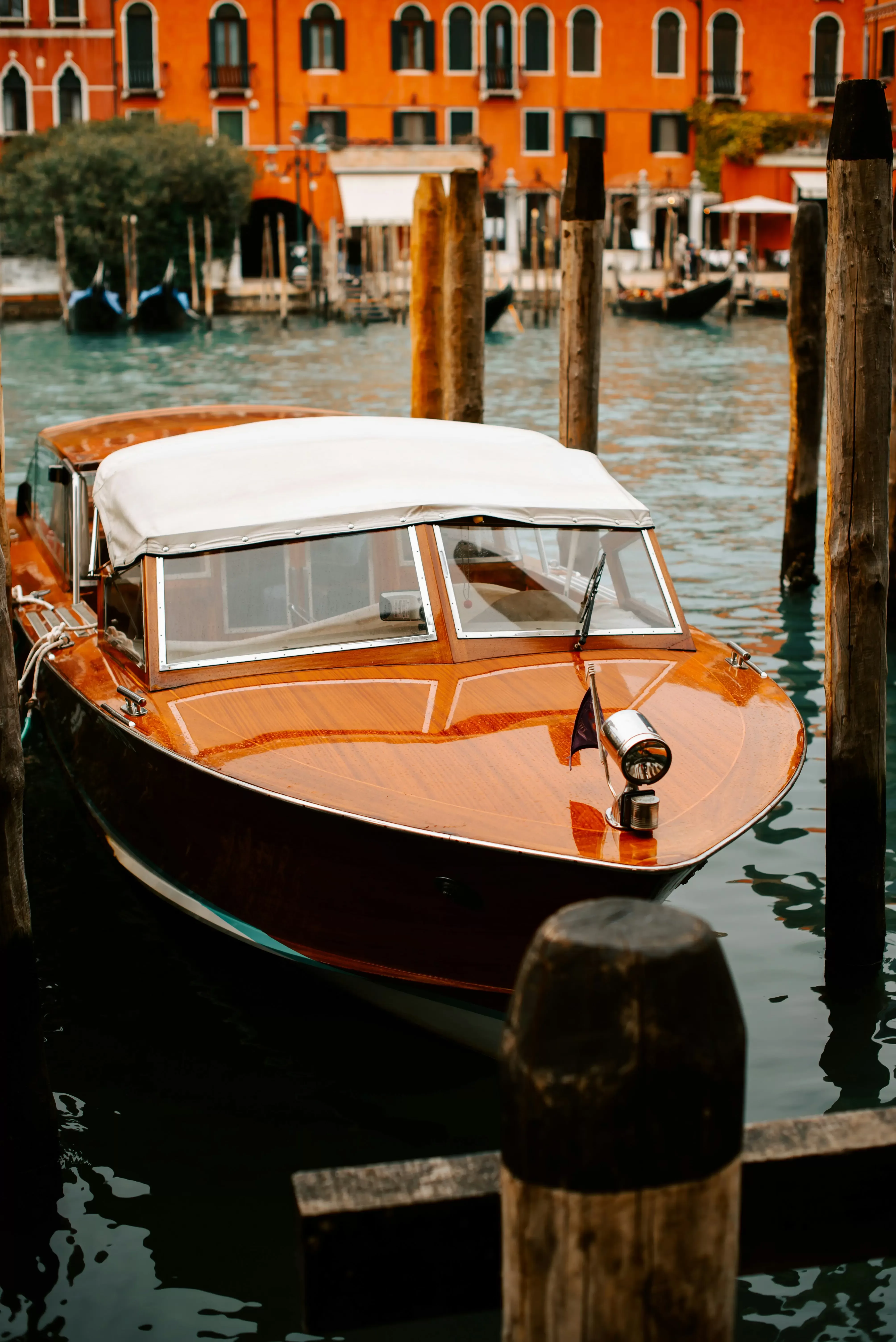 A wooden boat with a white canopy is docked between wooden posts on a canal. In the background, there is a vibrant orange building with arched windows and gondolas.
