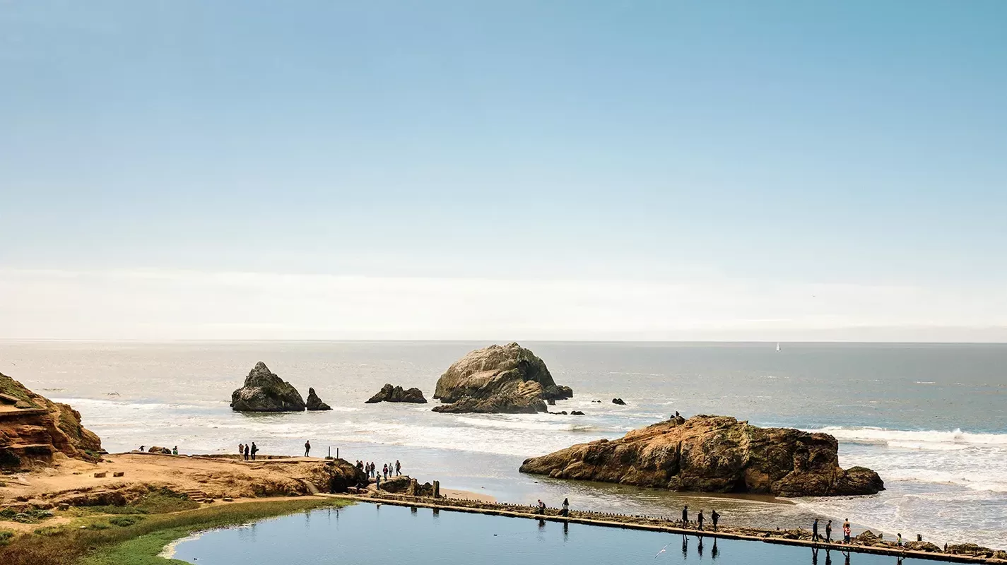 A scenic view of a rocky coastline with large boulders in the sea. People are walking along the edge of a clear, reflective pool adjacent to the ocean. The sky is clear and blue, creating a serene atmosphere.