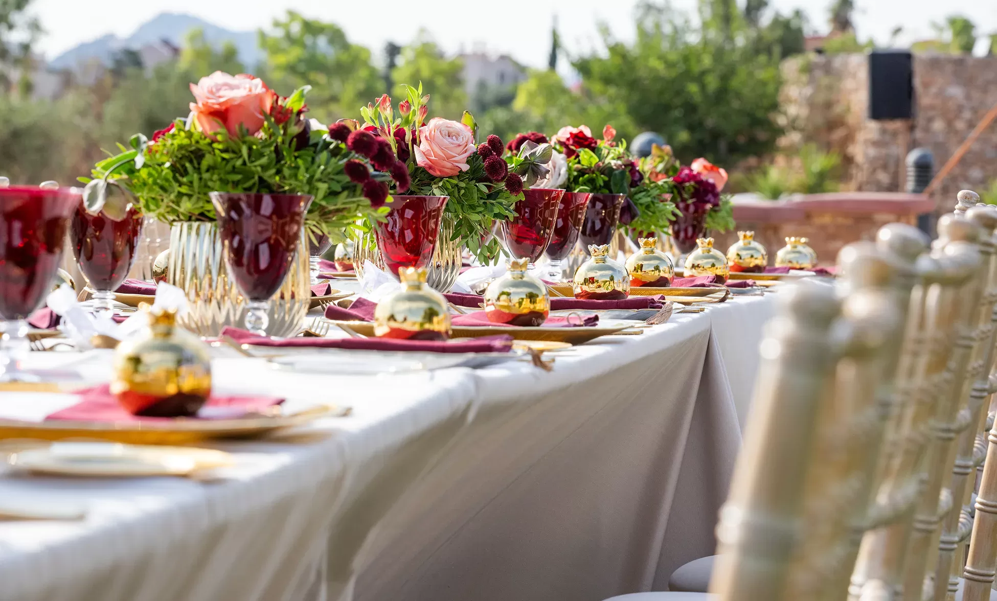An outdoor banquet table is elegantly set with gold and burgundy accents, red and pink floral centerpieces, gold utensils, and glassware, with greenery and trees visible in the background.