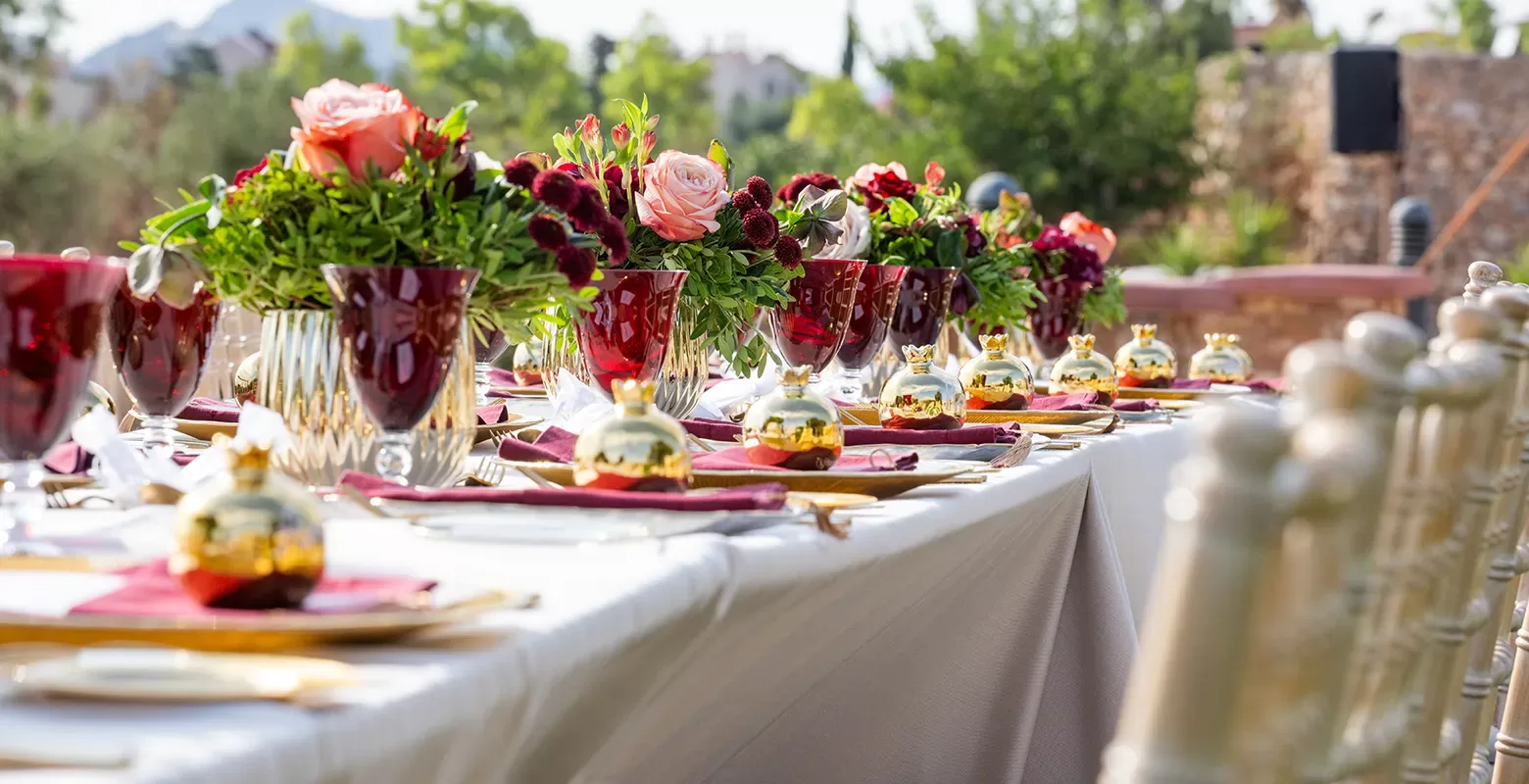 A long outdoor table is elegantly set with white linens, red and gold accents, floral centerpieces of roses and greenery, and small golden ornaments, surrounded by chairs in a lush garden setting.