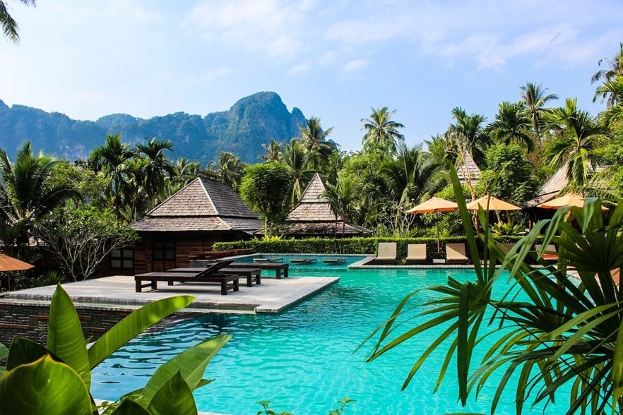 A tropical resort pool scene with wooden lounge chairs and umbrellas, surrounded by lush greenery and palm trees. In the background, mountains rise under a blue sky with scattered clouds.