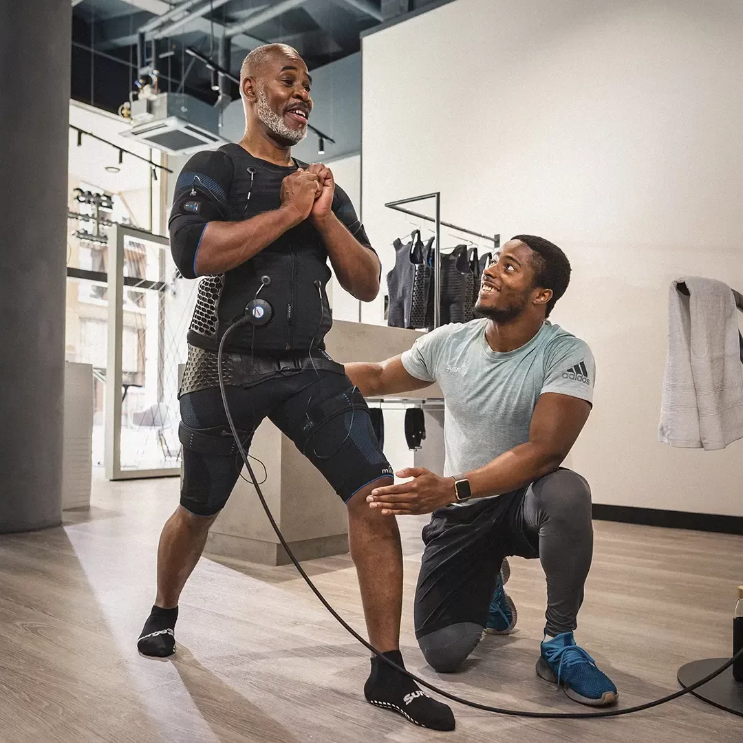 Man lunging in a gym wearing a EMS (electro-muscle stimulation) smiling at Personal Trainer