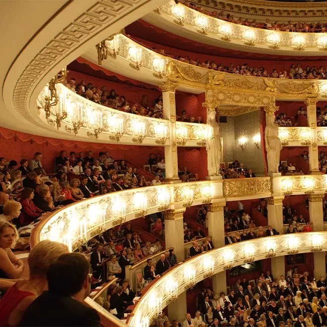 People sitting on tiered red theatre chairs overlooking gold balconies at Bavarian State Opera in Munich.