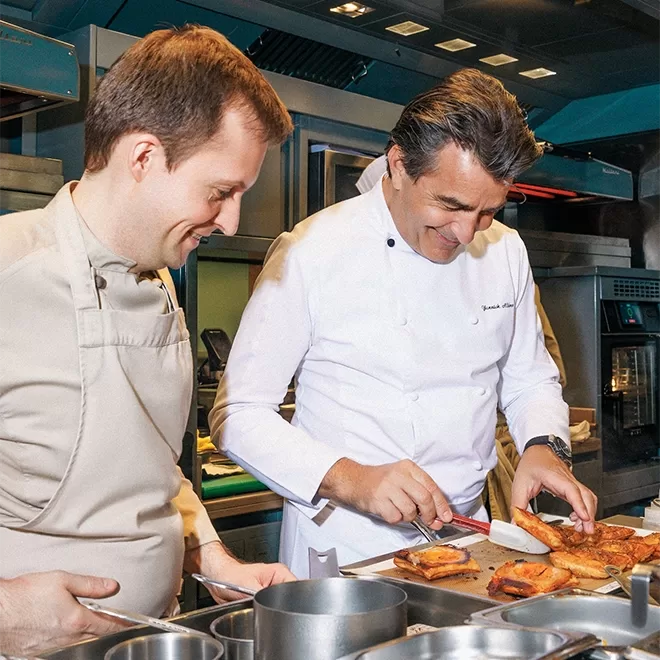 Two chefs in white uniforms smile while preparing food in a modern kitchen, with one chef slicing pastries on a tray and the other observing closely. Stainless steel pots and kitchen equipment are visible around them.