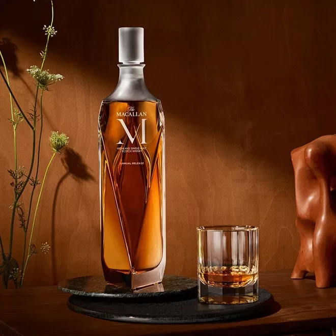 A bottle of The Macallan single malt whisky with an elegant, sculpted design is displayed next to a glass of whisky on a dark, round stone coaster against a warm brown background with soft lighting and floral accents.