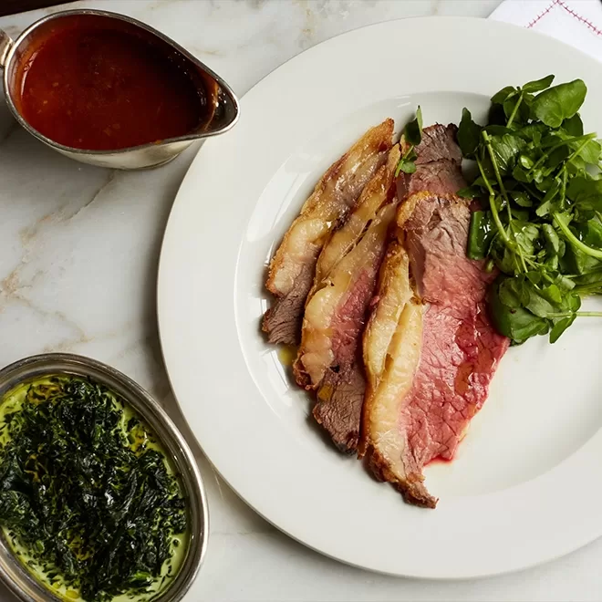 Three slices of roast beef with a garnish of watercress on a white plate, served with a side of green creamed spinach and a gravy boat of brown sauce on a marble surface.