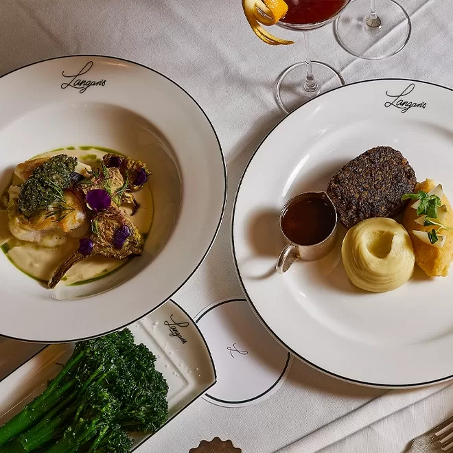 Two elegant plates of gourmet food on a white tablecloth: one with fish, sauce, and vegetables; the other with beef, mashed potatoes, sauce, and garnish. Broccolini and a cocktail are also visible. Both plates are labeled “Langans.”.