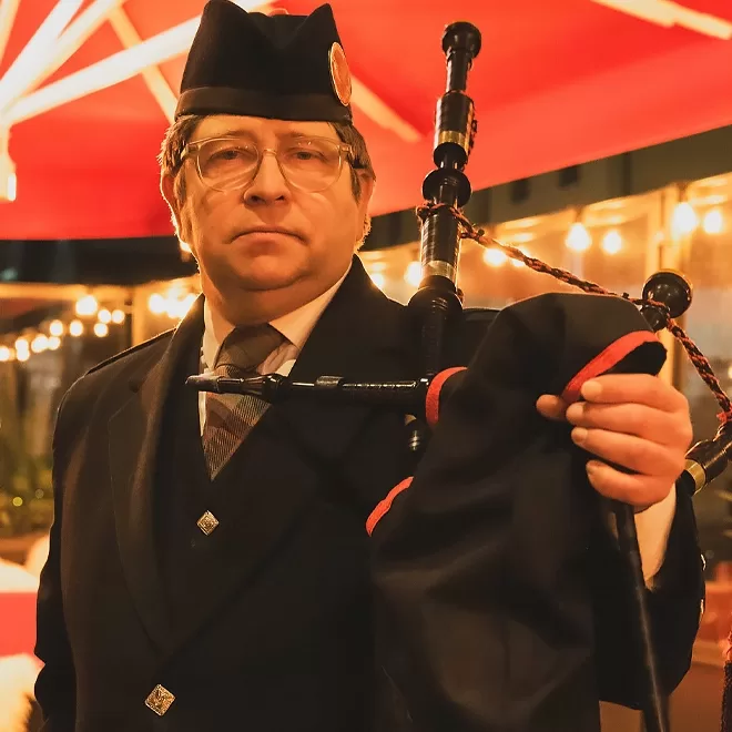 A man wearing a dark uniform and hat stands under bright lights, holding a bagpipe. He looks directly at the camera with a serious expression. Red and white umbrellas and string lights create a festive atmosphere in the background.