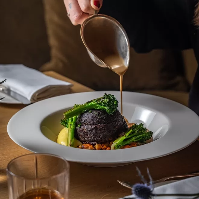 A person pours sauce over a gourmet dish featuring a dark round centerpiece, mashed potatoes, broccolini, and a vegetable medley, served in a white bowl on a wooden table.