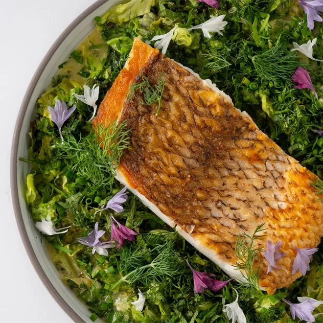 A cooked salmon fillet with crispy skin sits on a bed of leafy greens, fresh dill, and purple edible flowers on a white plate.