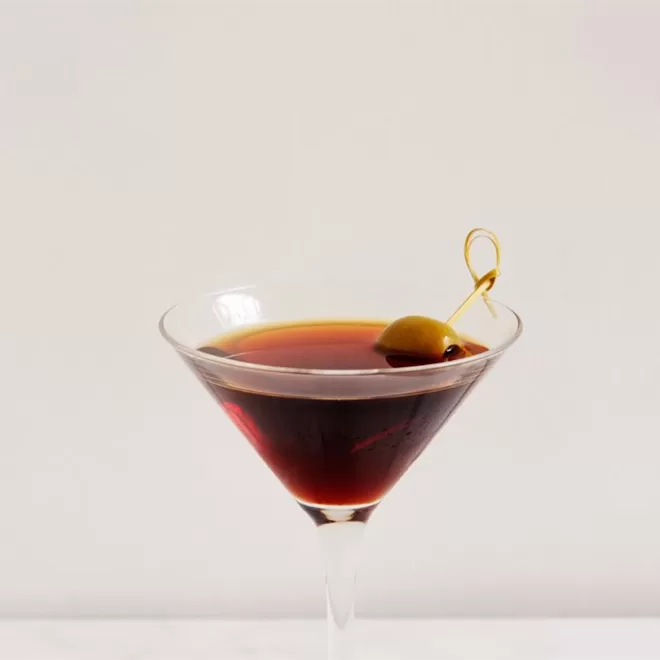 A classic martini glass filled with a dark brown cocktail, garnished with a green olive on a cocktail pick, set against a plain light background.