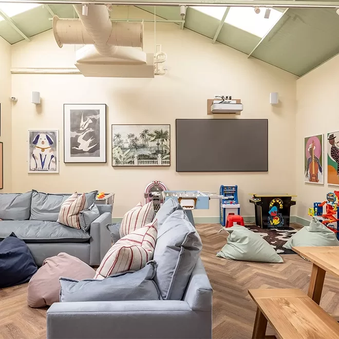 A cosy playroom with light blue sofas, striped pillows, bean bags, toys, and a play kitchen. Artworks and a blackboard hang on cream-colored walls beneath a high ceiling with skylights and exposed vents.