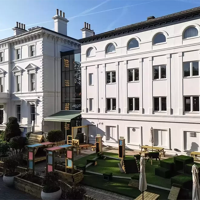 Outdoor courtyard with artificial grass, colorful play equipment, benches, and umbrellas surrounded by white, multi-story historic buildings under a clear blue sky.
