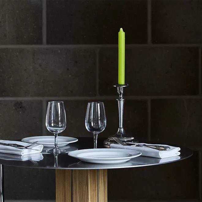 A round table set for two with white plates, folded napkins, cutlery, wine glasses, and a silver candlestick holding a tall green candle, against a dark brick wall.