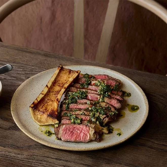 A plate with sliced medium-rare steak topped with green herb sauce, served alongside a roasted bone marrow piece on a wooden table.