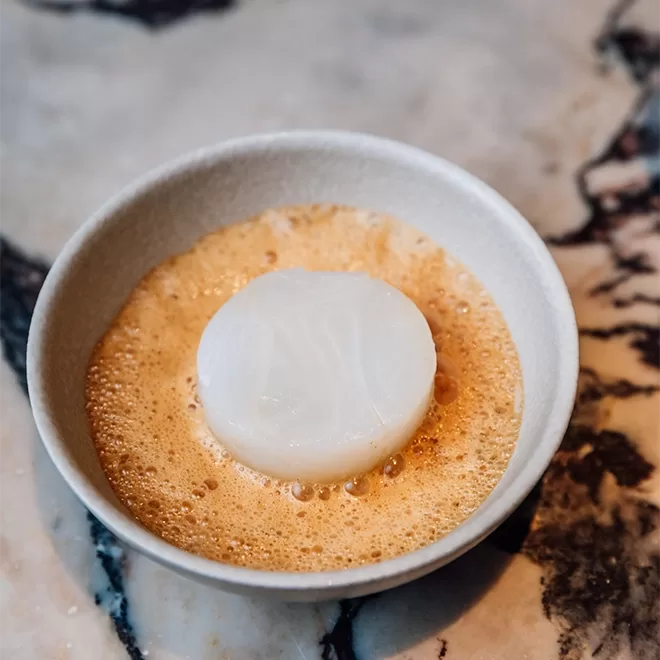 A white scallop sits atop a foamy, light brown sauce in a small, round, white bowl on a marbled surface.