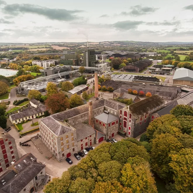 Aerial view of a historic stone distillery complex surrounded by trees, with industrial buildings and countryside in the background under a cloudy sky. Cars are parked near the main buildings.