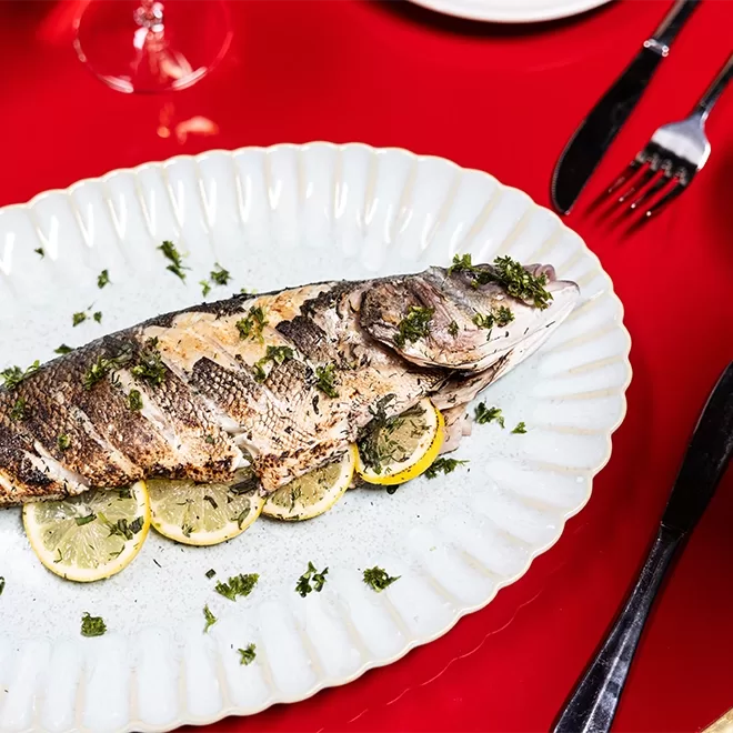 Grilled whole fish garnished with chopped herbs and lemon slices, served on a white oval plate on a red table, with a fork and knife next to the plate.
