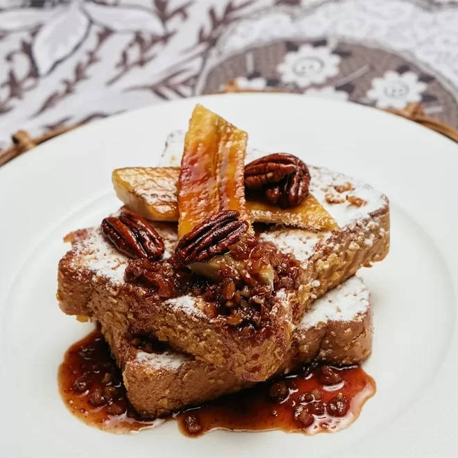Two thick slices of French toast topped with caramelized banana slices, pecans, powdered sugar, and drizzled with a rich, brown syrup on a white plate.