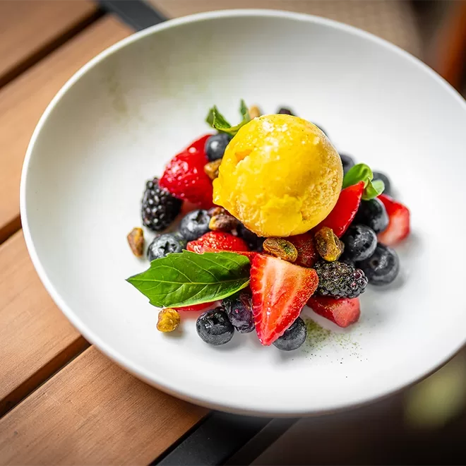 A white bowl with a scoop of yellow sorbet on top of fresh blueberries, strawberries, and blackberries, garnished with mint leaves and pistachios, placed on a wooden table.
