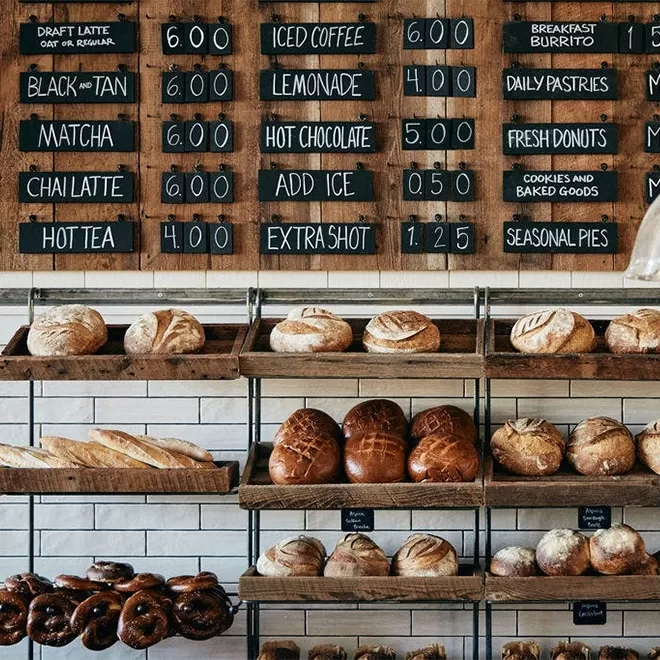 Shelves filled with various loaves of bread and pastries sit below a rustic menu board listing drinks and breakfast items, all with wooden and chalkboard decor in a cozy bakery setting.