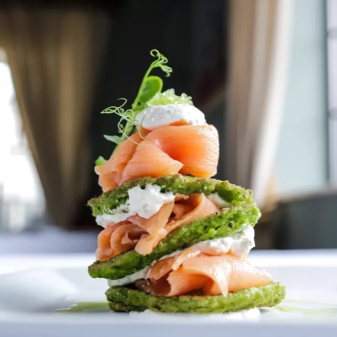 A gourmet stack of green pancakes layered with smoked salmon, cream, and garnished with herbs, presented on a white plate in a softly lit, elegant dining setting.