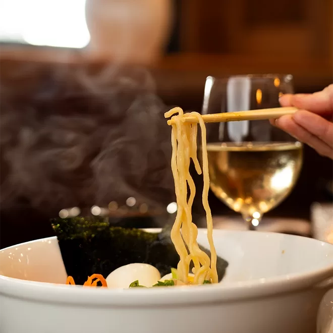 A hand holding chopsticks lifts steaming ramen noodles from a white bowl, with seaweed, vegetables, and a glass of white wine in the blurred background.