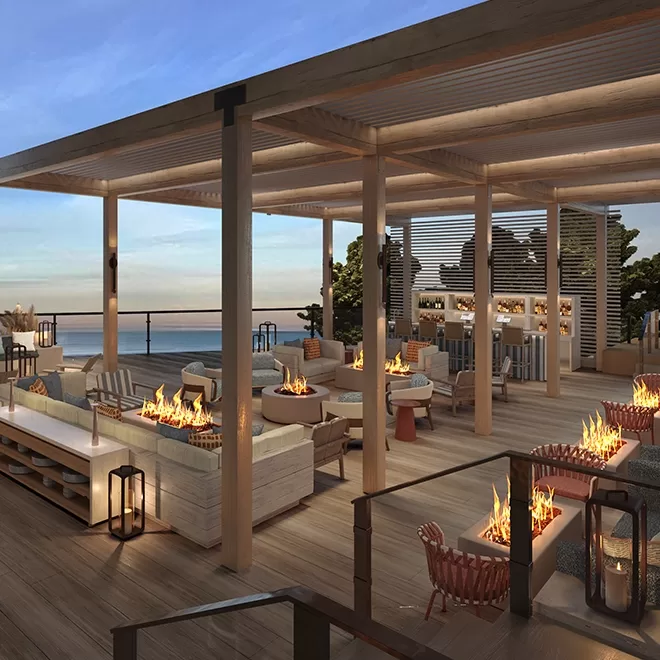 A modern outdoor rooftop lounge with wooden decking, cosy sofas, armchairs, and several fire pits. The area is covered by a pergola and overlooks a scenic ocean view at sunset.