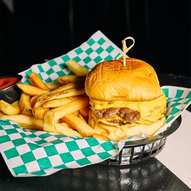 A cheeseburger with melted cheese and a beef patty is served in a basket with golden French fries on green-and-white checkered paper. A small cup of ketchup is on the side.