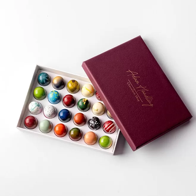 An open maroon box with 24 colorful, spherical chocolates arranged neatly in rows, each with unique artistic designs, on a white background.