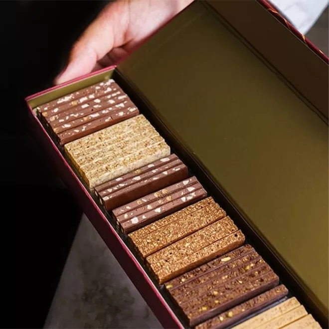 A hand holds an open box containing neatly arranged rows of rectangular chocolate and wafer biscuits in different shades of brown and beige.