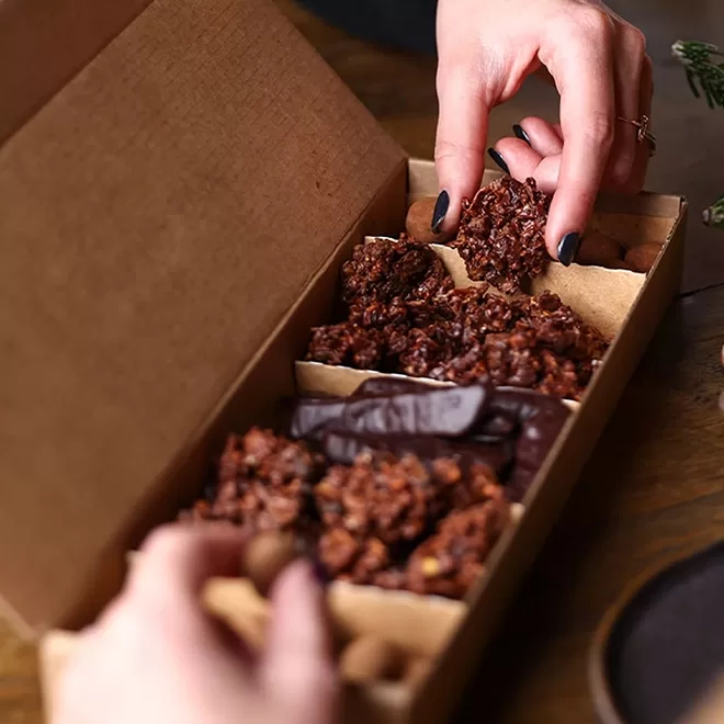 A person picks up a chocolate cluster from a brown cardboard box filled with assorted chocolates and treats, with another hand holding the box.