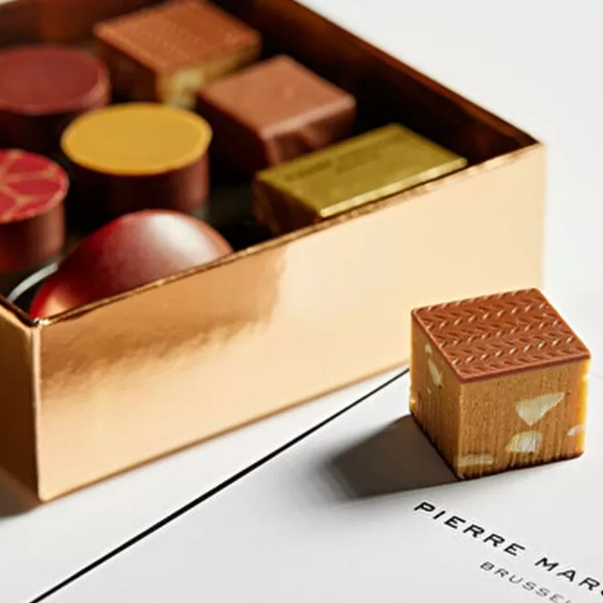 A close-up of assorted gourmet chocolates in a gold box, with one square chocolate with nuts placed outside on a white surface next to the words Pierre Marcolini Brussels.