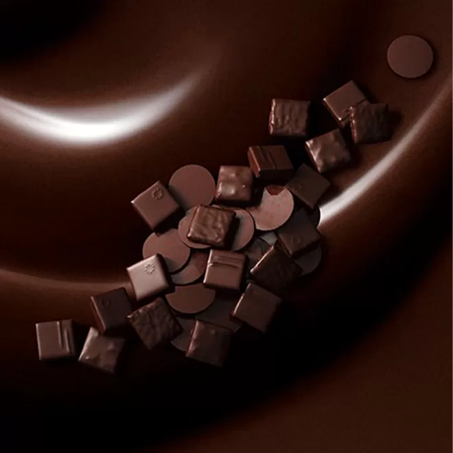 A close-up of dark chocolate pieces and round chocolate discs scattered on a smooth, shiny, swirling surface of melted chocolate.