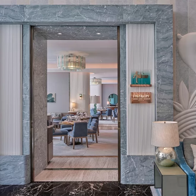A stylish restaurant entrance with a gray marble frame, a modern lamp on a side table, and a view of elegant dining tables and chairs inside under contemporary chandeliers.