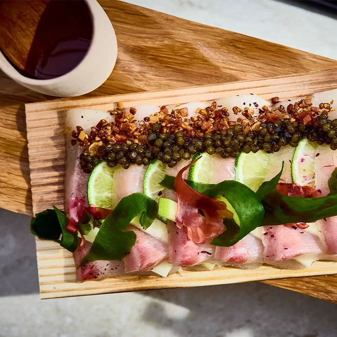 A wooden platter with neatly arranged sashimi slices, topped with lime wedges, caviar, and crispy bits, garnished with green leaves and pickled ginger. A small bowl of dark dipping sauce sits beside the platter.