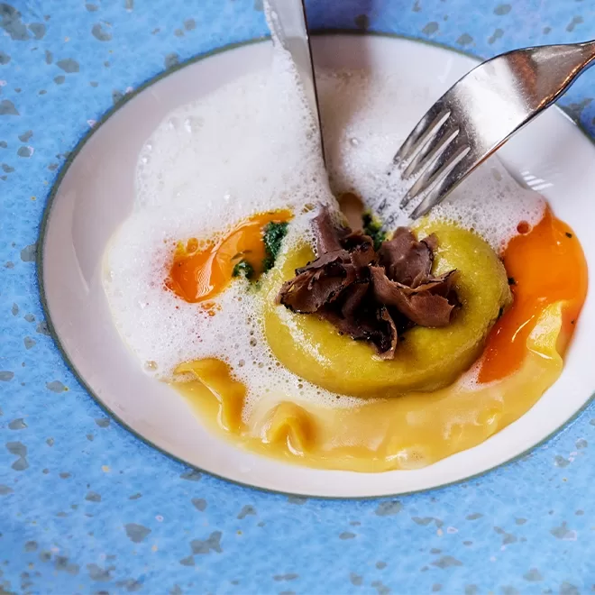A fork and knife cut into a round piece of stuffed pasta topped with shaved truffle, surrounded by egg yolk and white foam, on a white plate with a blue speckled rim.