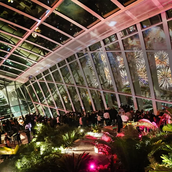 A crowd watches a fireworks display through the large slanted glass windows of a modern indoor garden at night, with colorful lights illuminating plants in the foreground.