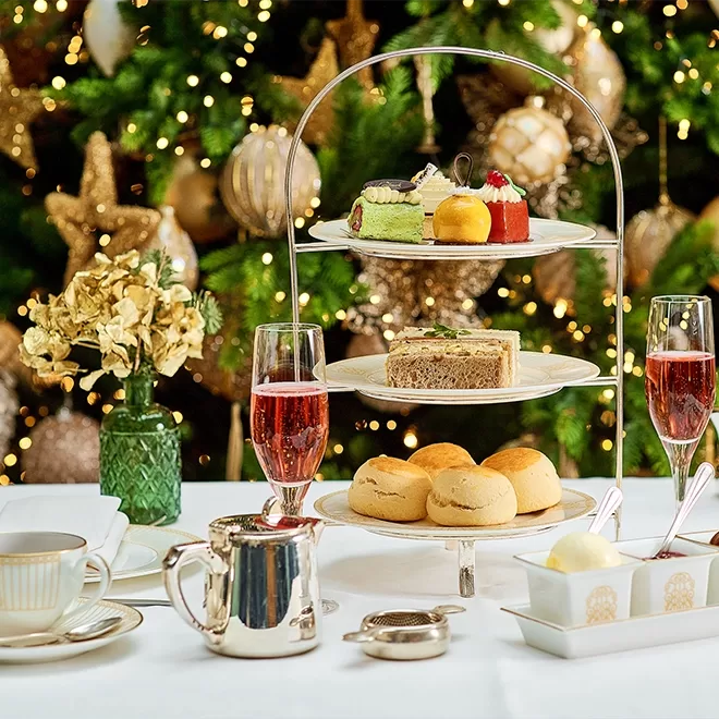 An elegant afternoon tea setup with sandwiches, pastries, and scones on a tiered stand, flanked by two glasses of pink tea, teacups, a teapot, and a vase of dried flowers, with a decorated Christmas tree in the background.