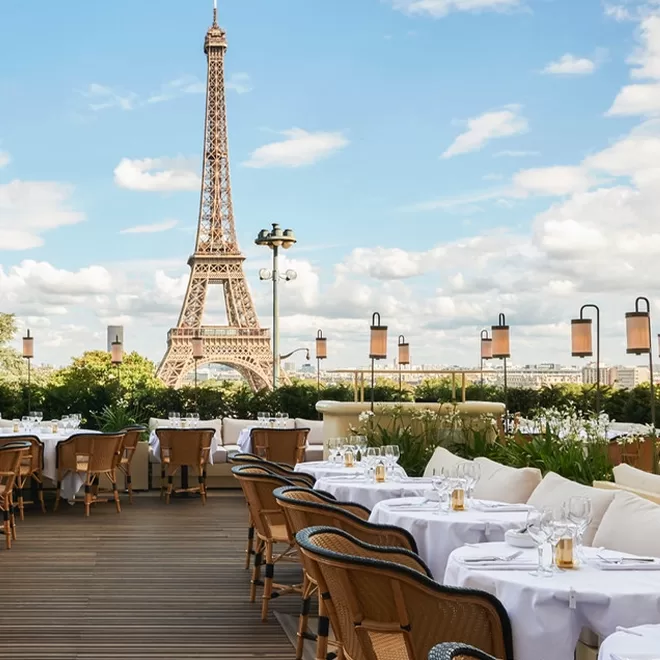 Outdoor restaurant terrace with elegant tables and chairs, set for dining, overlooking lush greenery and the Eiffel Tower under a partly cloudy blue sky in Paris.