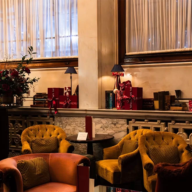 A cozy, elegant lounge with mustard-yellow and pink armchairs, marble-topped tables, books, red gift bags, and flower arrangements, softly lit by table lamps beneath tall windows with sheer curtains.