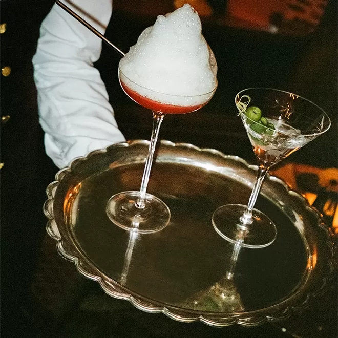 A person holding a silver tray with two cocktails: one with a foamy, frothy top and a straw, the other a clear drink garnished with green olives.