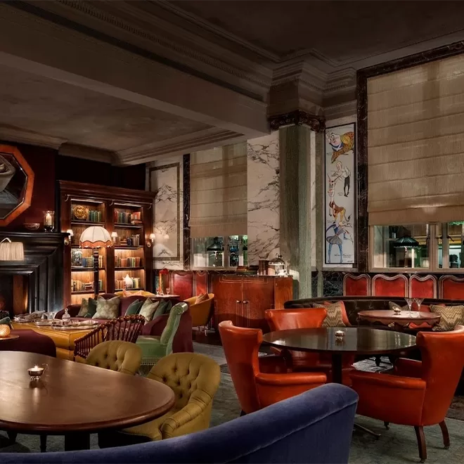 A stylish, dimly lit lounge with plush armchairs, round wooden tables, bookshelves, and a fireplace. Large windows with blinds and artistic wall panels add to the cozy, sophisticated ambiance.