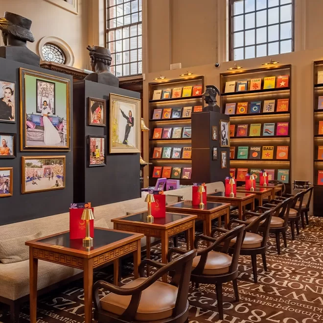 A stylish café with wooden tables and chairs, decorated with colorful bookshelves and framed art on the walls. Large windows let in natural light, and red menus are placed on each table.