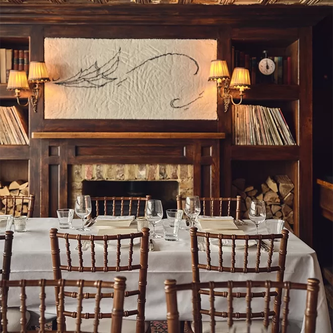 A cozy restaurant dining area with a wooden fireplace, bookshelves, stacked firewood, elegant table settings, and warm lighting from wall sconces.