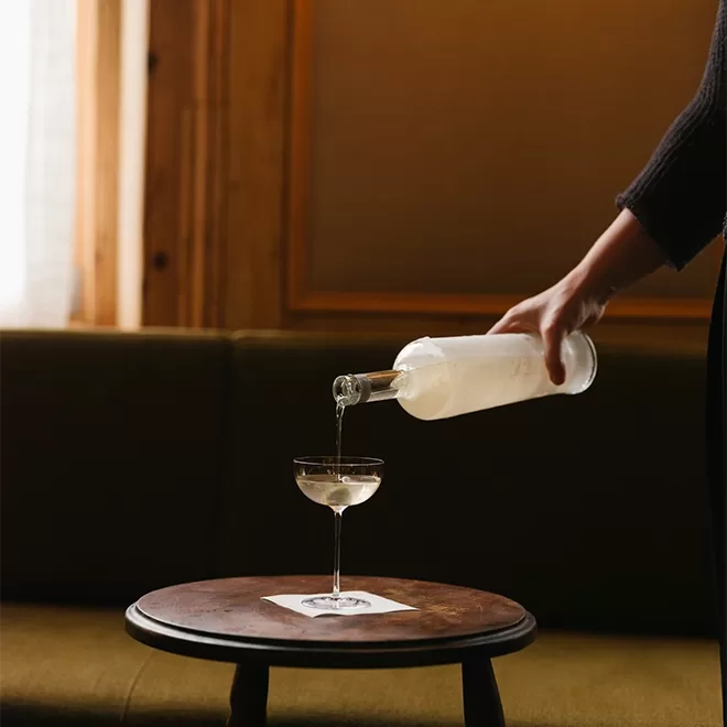 A person pours a clear liquid from a bottle into a cocktail glass on a small round wooden table, with a green sofa and wooden wall in the background.