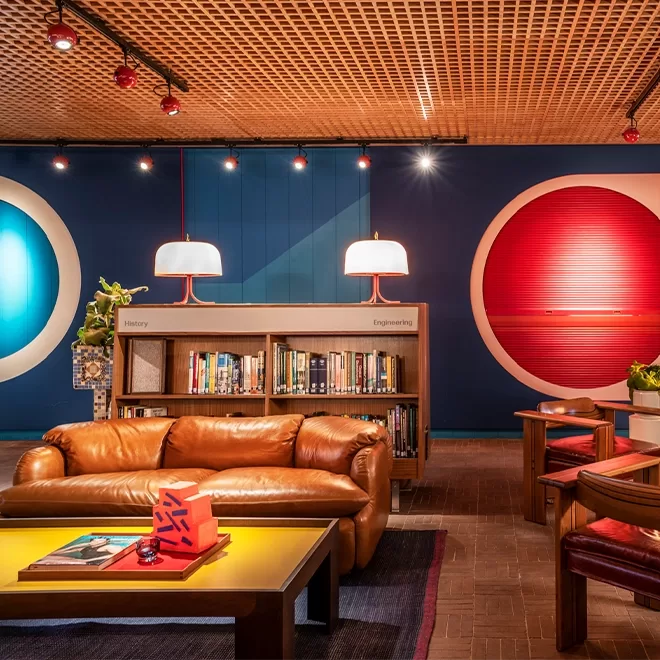 A modern library lounge with a tan leather sofa, yellow coffee table, and bookshelves. The background features bold blue and red circular wall designs and pendant lamps with white shades, creating a vibrant, inviting atmosphere.