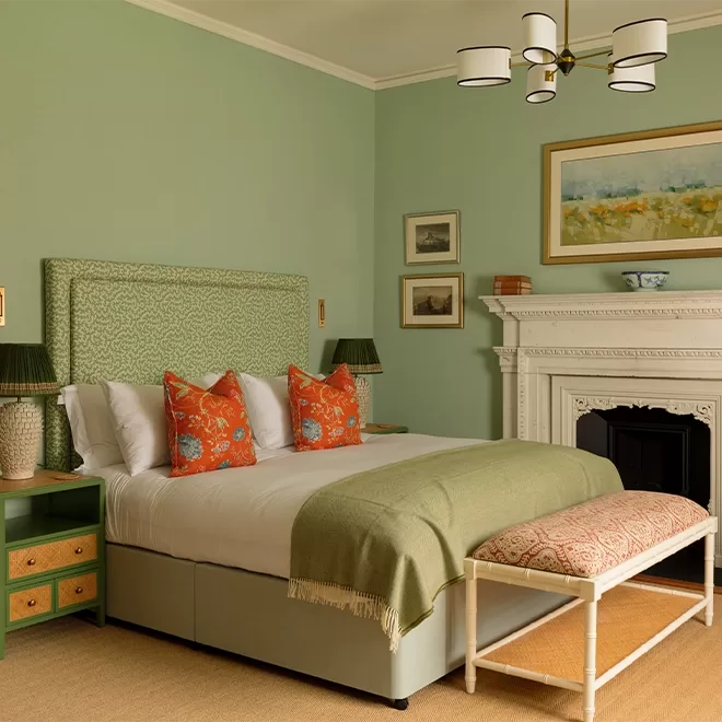 A cozy bedroom with green walls, a bed with a green headboard, white bedding, and orange floral pillows. There’s a green blanket, a patterned bench at the foot, green nightstands, lamps, and a white fireplace with wall art above.