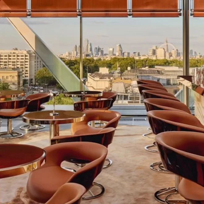 A modern lounge with round brown chairs and small tables, featuring floor-to-ceiling windows offering a panoramic city view with skyscrapers and landmarks in the background.